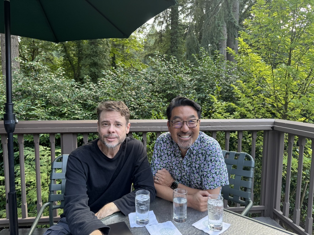 Steve Yegge and Gene Kim seated outdoors on a tree-lined deck during a co-author working session
